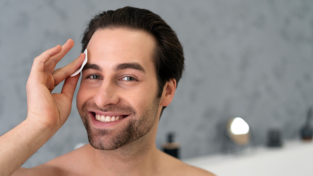 Homem sorrindo passando um disco de algodão no rosto após fazer procedimentos estéticos para homens.