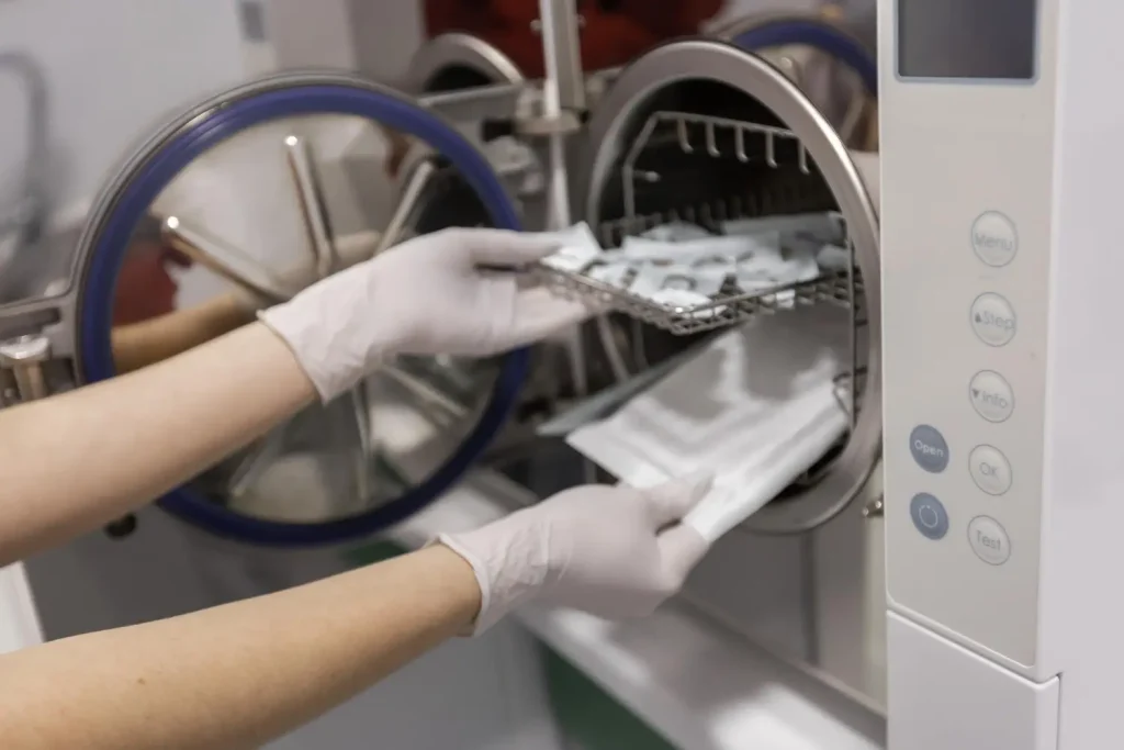 Mãos usando luvas para retirar bandeja com utensílios esterilizados do autoclave, ilustrando o funcionamento do autoclave em clínica odontológica.