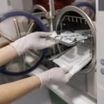 Mãos usando luvas para retirar bandeja com utensílios esterilizados do autoclave, ilustrando o funcionamento do autoclave em clínica odontológica.